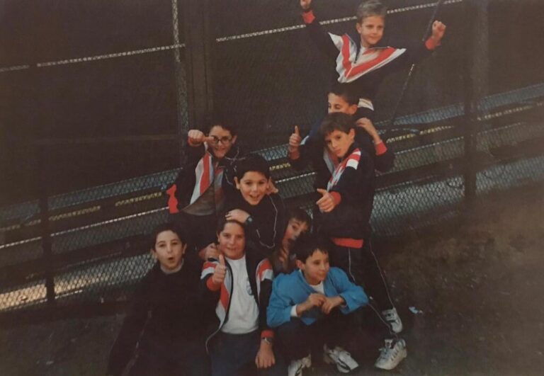 Grupo de niños con chándales a juego posando sonrientes junto a una valla metálica y haciendo gestos de victoria