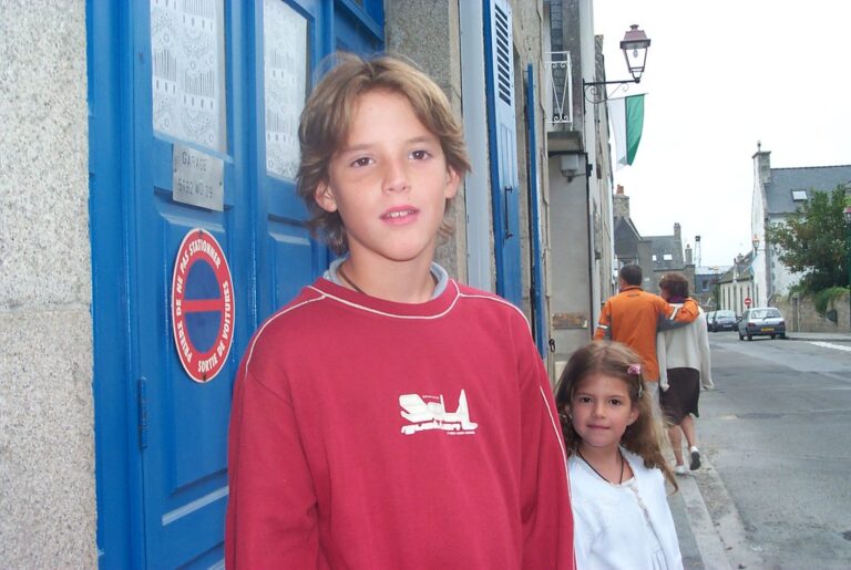 Marcos Pou de niño, con sudadera roja, junto a una niña en una calle con puerta azul y pareja al fondo