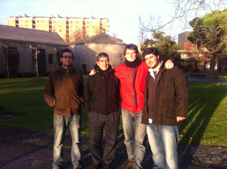 Cuatro hombres abrigados posan juntos en un parque, con una iglesia de piedra y bloques de pisos al fondo