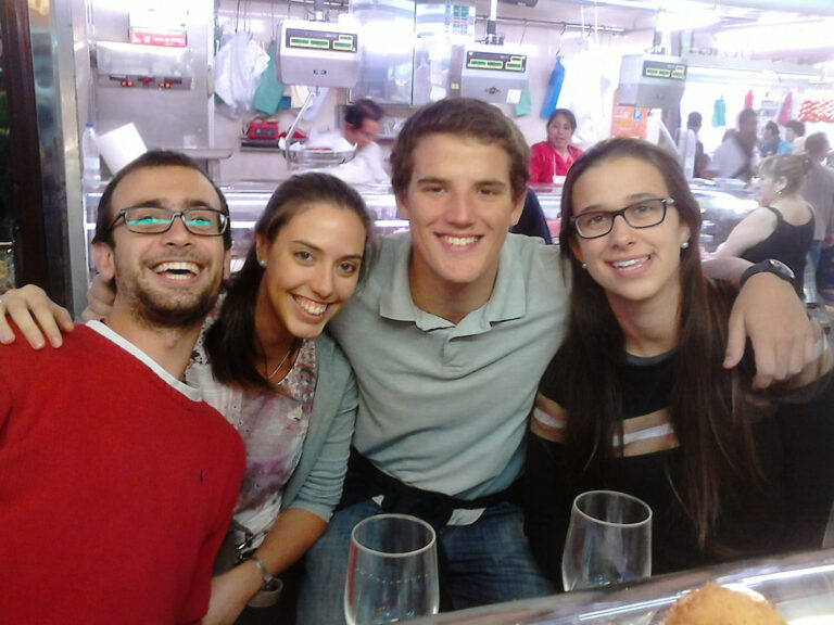 Marcos Pou abrazado a dos chicas y un chico, sonriendo ante la barra de un bar dentro de un mercado
