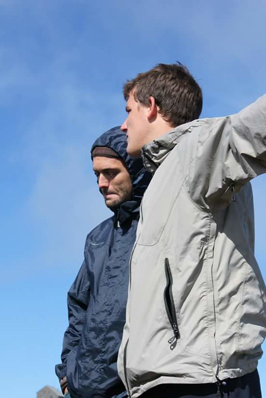 Marcos Pou, con cortavientos claro, señala el horizonte junto a otro joven con chubasquero azul bajo un cielo despejado