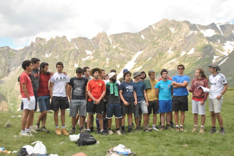 Grupo de jóvenes de pie en un prado de montaña, escuchando a un compañero que lee unas hojas
