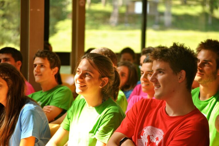 Grupo de jóvenes con camisetas de colores sonriendo y escuchando atentamente durante una charla en una sala con ventanales al campo