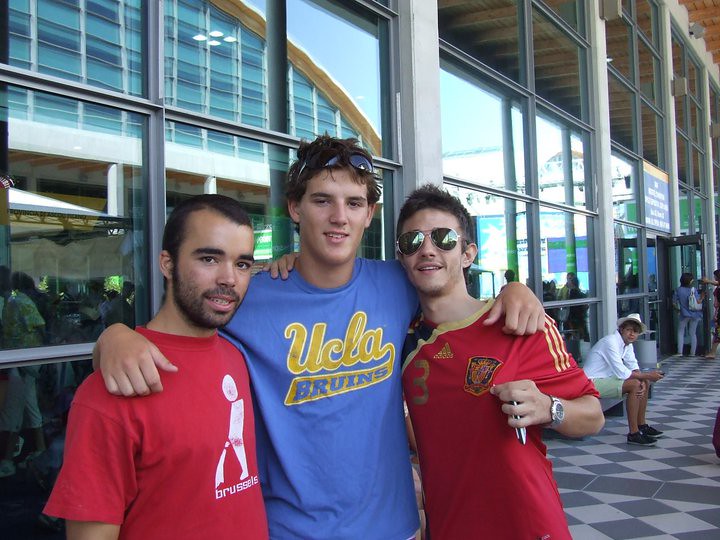 Tres jóvenes abrazados y sonrientes ante un edificio acristalado, con camisetas de Bruselas, UCLA y de la selección española