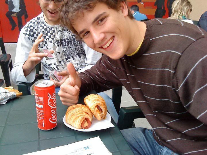 Marcos Pou sonriente levanta el pulgar ante un plato de bollos y una lata de refresco, junto a un amigo con cartas en la mano