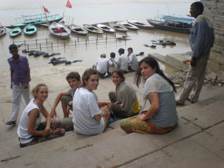 Un grupo de jóvenes sentados en las escaleras de un río en la India, con barcas y búfalos en el agua al fondo
