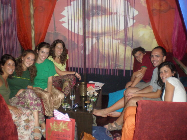 Grupo de jóvenes sentados en sofás y cojines de un local con cortinas rojas, sonriendo alrededor de una mesa baja