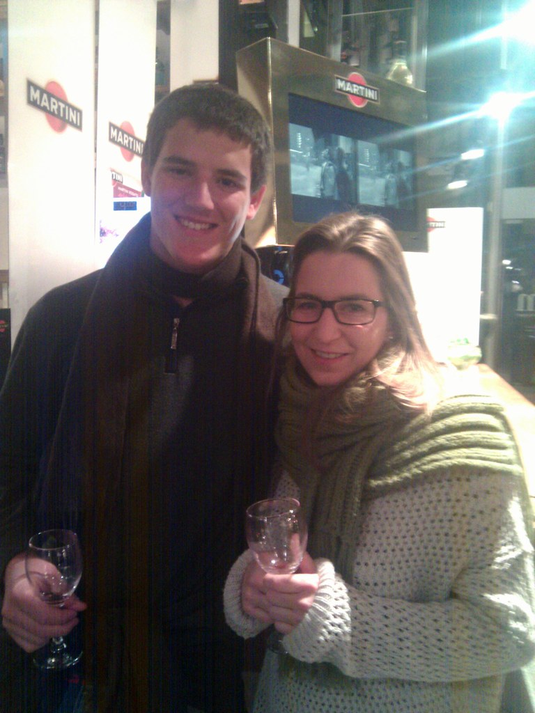 Marcos Pou, con bufanda y copa en la mano, sonríe junto a una joven con gafas en un bar con carteles de Martini