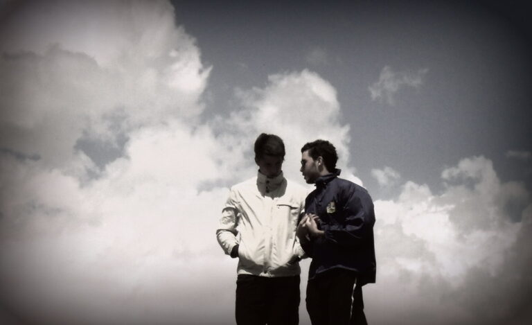 Dos jóvenes con cortavientos conversan al aire libre recortados contra un cielo de grandes nubes blancas