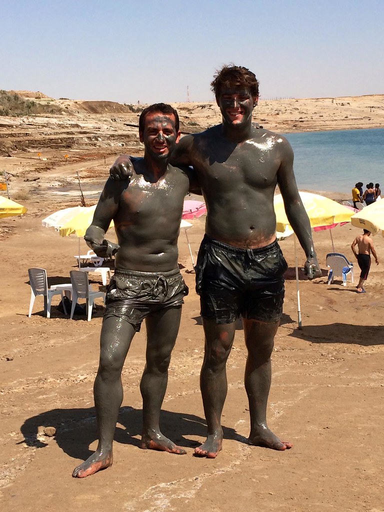 Dos jóvenes sonrientes cubiertos de barro posan abrazados en la orilla del Mar Muerto, entre sombrillas y sillas de playa