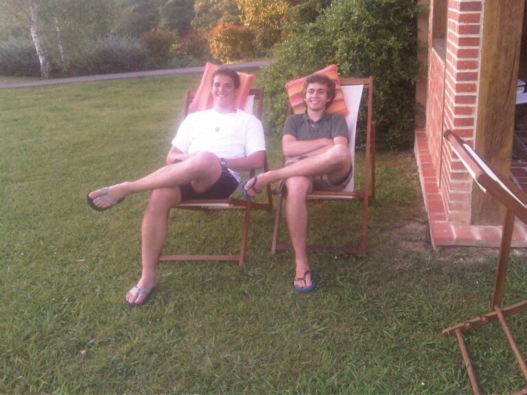 Dos jóvenes sonrientes sentados en tumbonas de madera sobre el césped de un jardín, junto a una casa de ladrillo