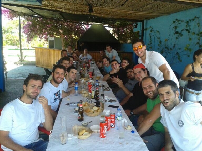 Un grupo numeroso de jóvenes sentados a una larga mesa alargada en una comida al aire libre bajo un porche