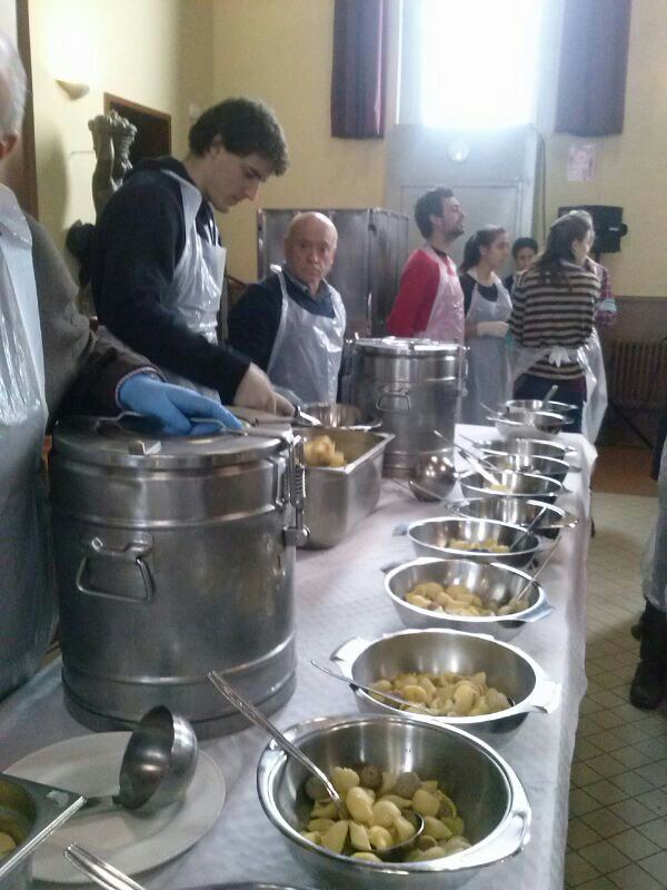 Grupo de voluntarios con delantal sirviendo comida en un comedor social, junto a ollas y fuentes de patatas sobre una mesa larga