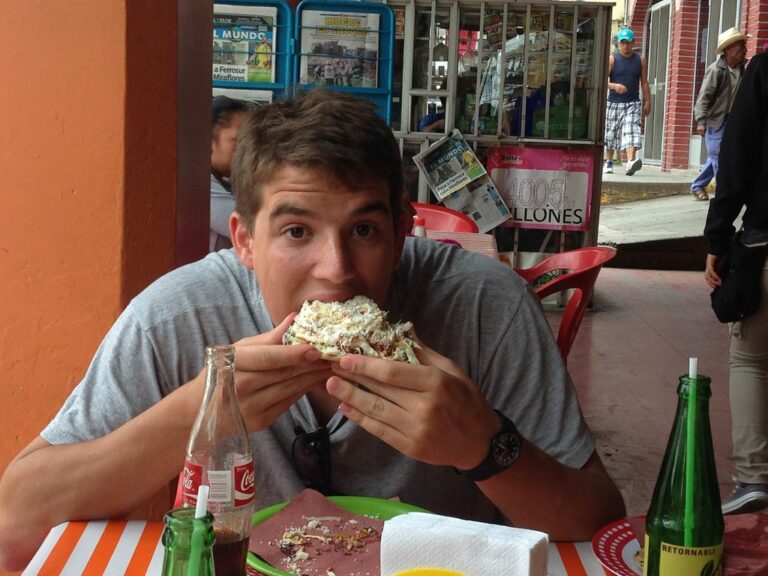 Marcos Pou comiendo un bocadillo en la terraza de un puesto callejero, con botellas sobre la mesa y gente al fondo