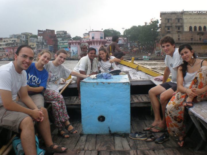 Grupo de jóvenes sentados en una barca de madera junto a un barquero, con edificios y el río al fondo durante un viaje