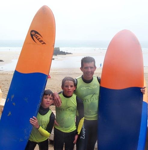 Dos niños y un hombre con licras verdes de surf posan entre tablas naranjas y azules en una playa