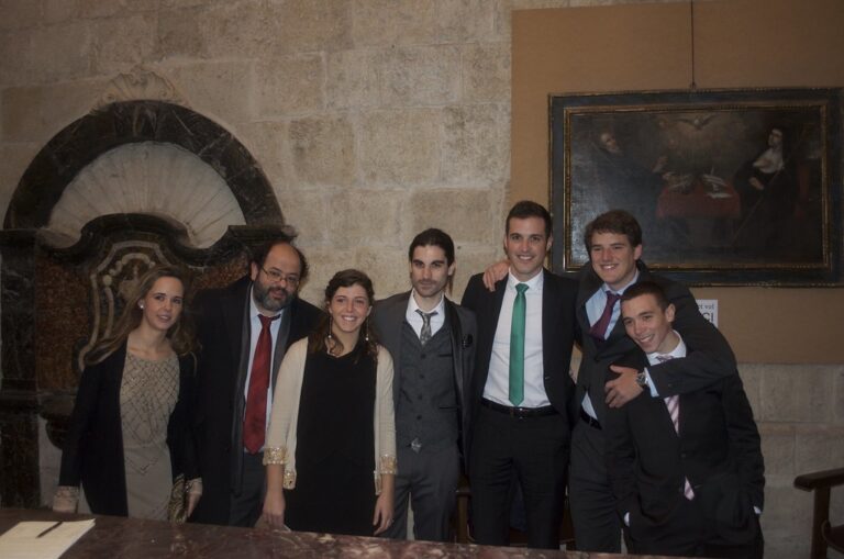 Grupo de jóvenes vestidos de traje y de fiesta posando juntos y sonrientes en una sala de piedra con un cuadro antiguo