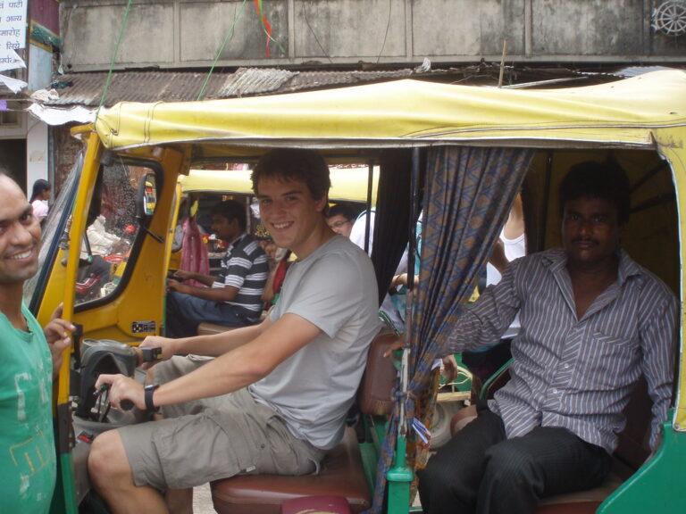 Marcos Pou sonriente al volante de un mototaxi en la India, acompañado por dos hombres locales en una calle concurrida