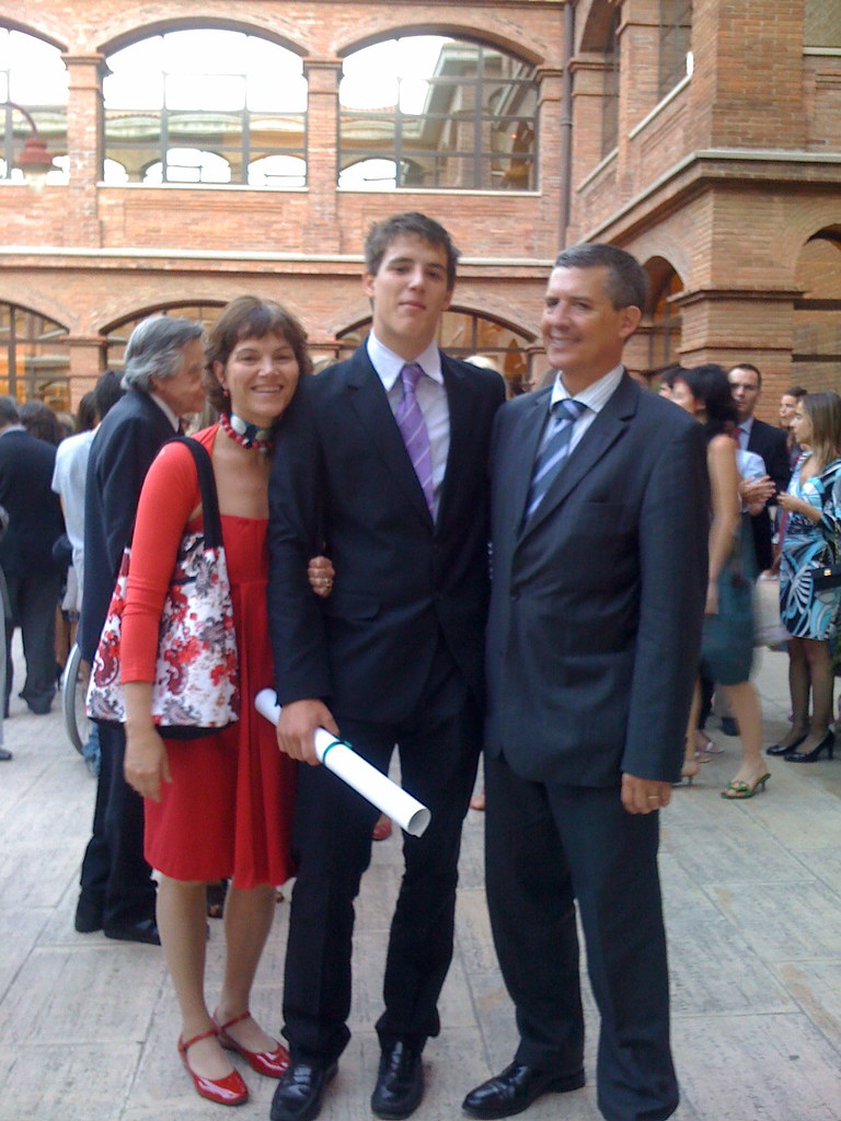 Marcos Pou, con traje y un diploma enrollado en la mano, posa entre sus padres en el patio de un edificio de ladrillo tras la graduación