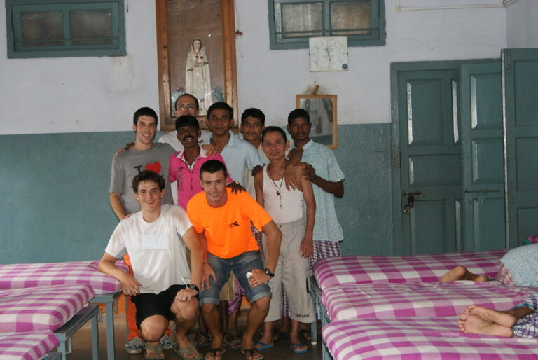 Grupo de jóvenes voluntarios posando con varios hombres en una sala con camas de cuadros y una imagen de la Virgen en la pared