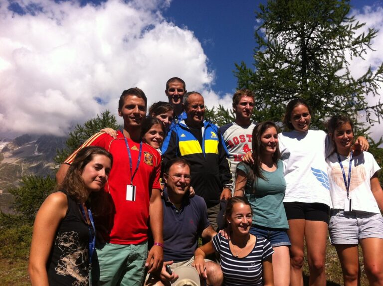 Grupo de jóvenes y dos adultos posando juntos en la montaña, con cumbres nevadas y cielo de nubes al fondo