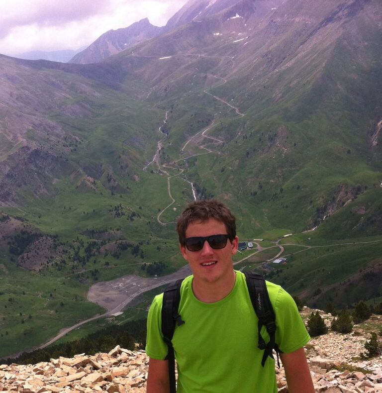 Marcos Pou, con camiseta verde, gafas de sol y mochila, sonríe en la cima de una montaña con un valle verde al fondo