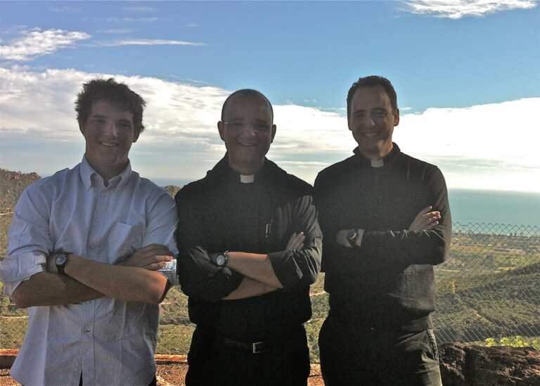 Marcos Pou junto a dos sacerdotes, sonrientes y con los brazos cruzados, en un mirador con el mar al fondo