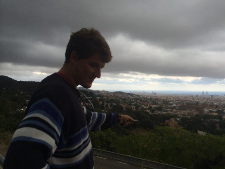 Marcos Pou, con jersey de rayas, sonríe en un mirador de la sierra con Barcelona y el mar al fondo bajo un cielo nublado