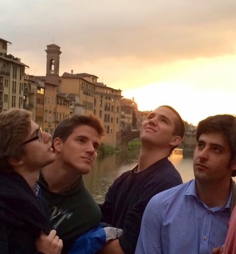 Cuatro jóvenes mirando hacia arriba junto a un río al atardecer, con edificios y un campanario al fondo