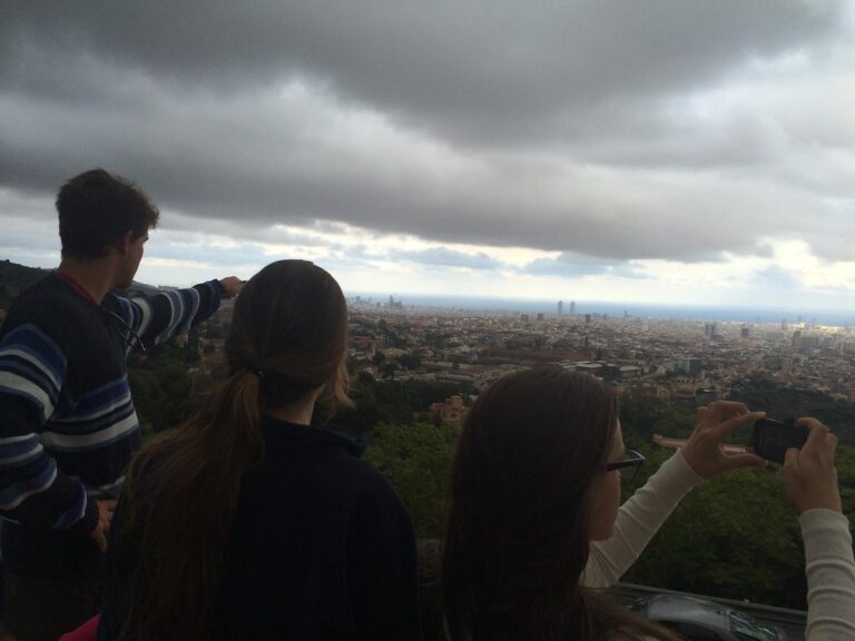 Marcos Pou señala el horizonte junto a dos jóvenes que miran y fotografían la ciudad de Barcelona desde un mirador con cielo nublado