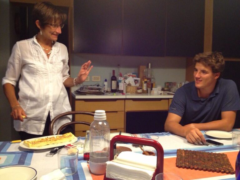 Marcos Pou sentado a la mesa de una cocina, sonriendo mientras una mujer de pie conversa con él durante una cena familiar