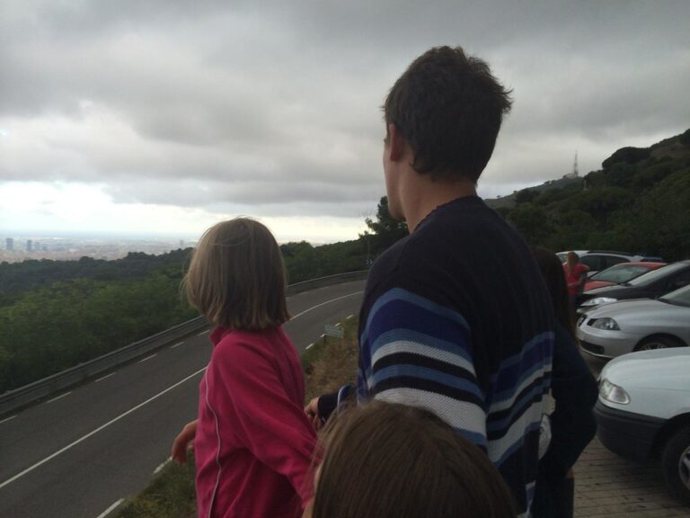 Marcos Pou, de espaldas, con unos niños en un mirador de la montaña contemplando la ciudad bajo un cielo nublado