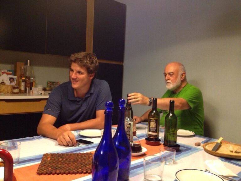 Marcos Pou sonriente sentado a la mesa de una cocina junto a un hombre mayor que sirve vino