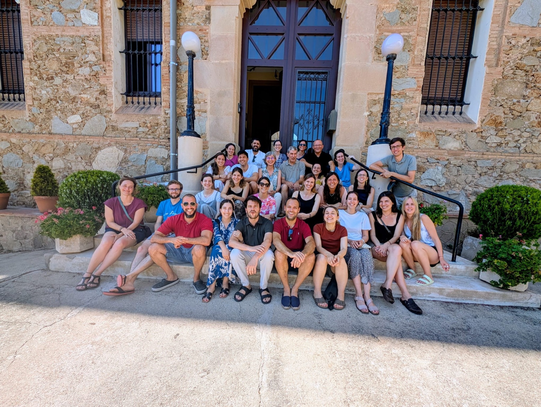 Grup d'amics de l'Associació Amics de Marcos Pou asseguts a l'escala de pedra d'una casa, a l'estiu